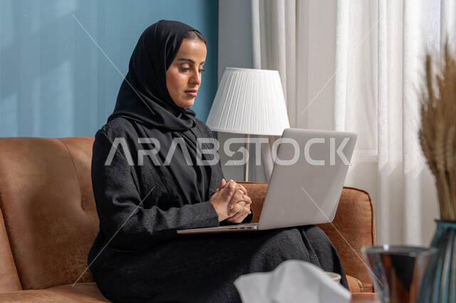 العمل على الحاسوب المحمول في صالة المنزل، ادارة وانجاز المهام باستخدام جهاز تقني حديث ومتطور, مهنة ووظيفة مكتبية، إمرأة عربية خليجية سعودية ترتدي الحجاب والعباءة السوداء تجلس على الكنبة