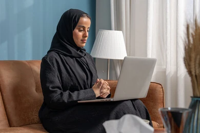 العمل على الحاسوب المحمول في صالة المنزل، ادارة وانجاز المهام باستخدام جهاز تقني حديث ومتطور, مهنة ووظيفة مكتبية، إمرأة عربية خليجية سعودية ترتدي الحجاب والعباءة السوداء تجلس على الكنبة
