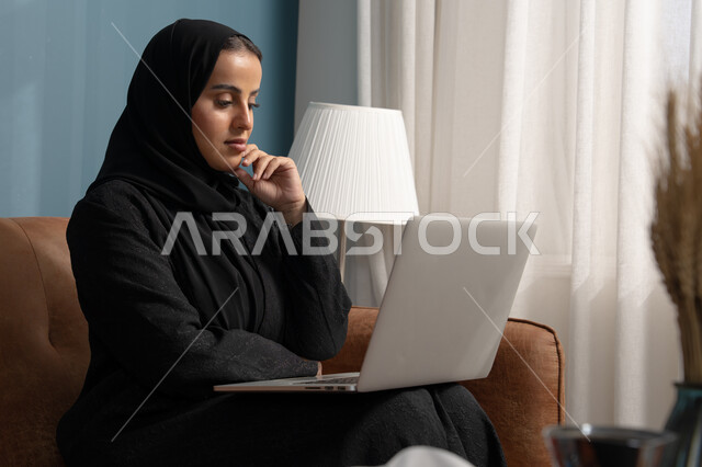 Working on a laptop in the living room, managing and completing tasks using a modern and advanced technical device, Saudi women's professions and jobs, a Saudi Arabian Gulf woman wearing a hijab and a black abaya sitting on the sofa