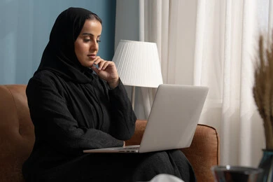 العمل على الحاسوب المحمول في صالة المنزل، ادارة وانجاز المهام باستخدام جهاز تقني حديث ومتطور, مهن ووظائف نسائية سعودية، إمرأة عربية خليجية سعودية ترتدي الحجاب والعباءة السوداء تجلس على الكنبة