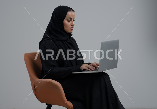مهنة ووظيفة مكتبية، ادارة وانجاز المهام باستخدام جهاز تقني حديث ومتطور, بورتريه لإمرأة عربية خليجية سعودية ترتدي الحجاب والعباءة السوداء تجلس الكرسي وتعمل على الحاسوب المحمول, خلفية رمادية