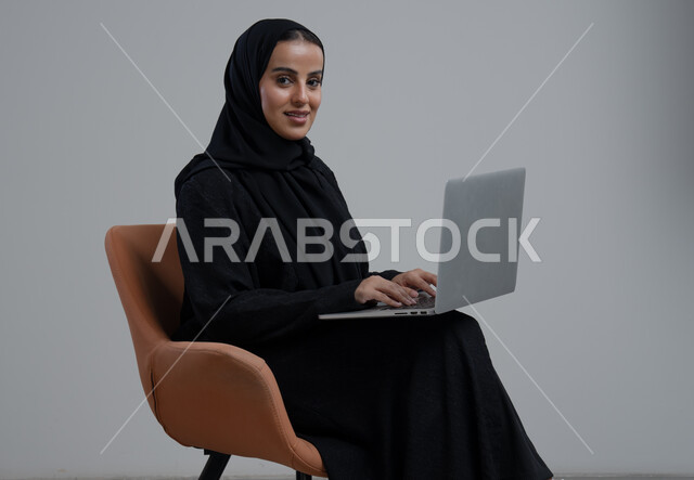 مهنة ووظيفة مكتبية، ادارة وانجاز المهام باستخدام جهاز تقني حديث ومتطور, بورتريه لإمرأة عربية خليجية سعودية ترتدي الحجاب والعباءة السوداء تجلس الكرسي وتعمل على الحاسوب المحمول, خلفية رمادية