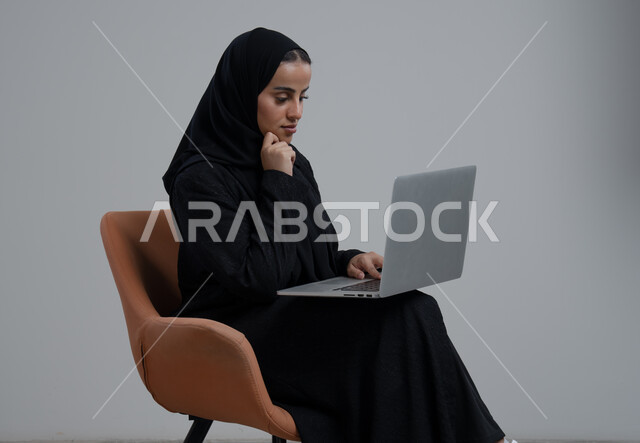 مهنة ووظيفة مكتبية، ادارة وانجاز المهام باستخدام جهاز تقني حديث ومتطور, بورتريه لإمرأة عربية خليجية سعودية ترتدي الحجاب والعباءة السوداء تجلس الكرسي وتعمل على الحاسوب المحمول, خلفية رمادية