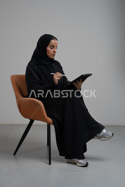 انجاز وادارة المهام باستخدام جهاز تقني حديث ومتطور, مهنة ووظيفة مكتبية، بورتريه لإمرأة عربية خليجية سعودية مبتسمة ترتدي الحجاب والعباءة تجلس على الكرسي و تعمل على جهاز التابلت, خلفية رمادية
