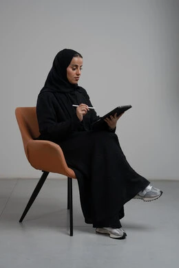 انجاز وادارة المهام باستخدام جهاز تقني حديث ومتطور, مهنة ووظيفة مكتبية، بورتريه لإمرأة عربية خليجية سعودية مبتسمة ترتدي الحجاب والعباءة تجلس على الكرسي و تعمل على جهاز التابلت, خلفية رمادية