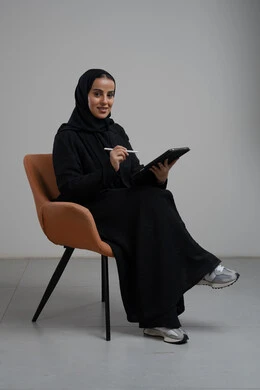 Accomplishing and managing tasks using a modern and advanced technical device, office profession and job, portrait of a smiling Saudi Arabian Gulf woman wearing a hijab and abaya sitting on a chair and working on a tablet, gray background