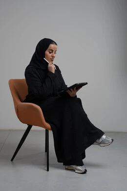 انجاز وادارة المهام باستخدام جهاز تقني حديث ومتطور, مهنة ووظيفة مكتبية، بورتريه لإمرأة عربية خليجية سعودية مبتسمة ترتدي الحجاب والعباءة تجلس على الكرسي و تعمل على جهاز التابلت, خلفية رمادية