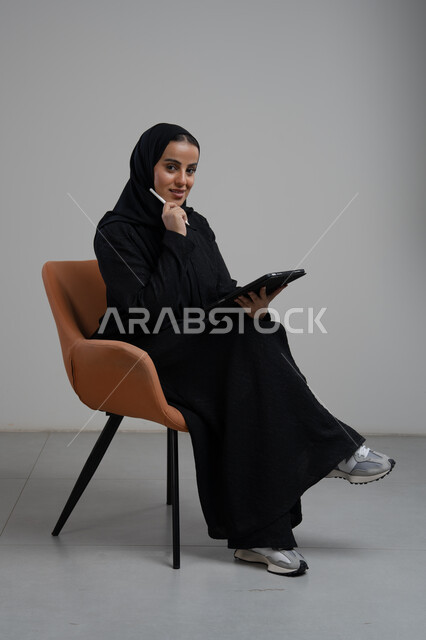 Accomplishing and managing tasks using a modern and advanced technical device, office profession and job, portrait of a smiling Saudi Arabian Gulf woman wearing a hijab and abaya sitting on a chair and working on a tablet, gray background