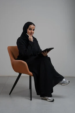 انجاز وادارة المهام باستخدام جهاز تقني حديث ومتطور, مهنة ووظيفة مكتبية، بورتريه لإمرأة عربية خليجية سعودية مبتسمة ترتدي الحجاب والعباءة تجلس على الكرسي و تعمل على جهاز التابلت, خلفية رمادية