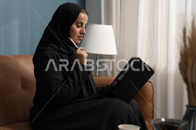 مهنة ووظيفة مكتبية، انجاز وادارة المهام باستخدام جهاز تقني حديث ومتطور, إمرأة عربية خليجية سعودية مبتسمة ترتدي الحجاب والعباءة وتجلس على الكنبة تعمل على جهاز التابلت في صالة المنزل