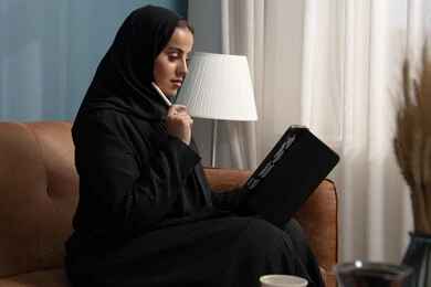 مهنة ووظيفة مكتبية، انجاز وادارة المهام باستخدام جهاز تقني حديث ومتطور, إمرأة عربية خليجية سعودية مبتسمة ترتدي الحجاب والعباءة وتجلس على الكنبة تعمل على جهاز التابلت في صالة المنزل
