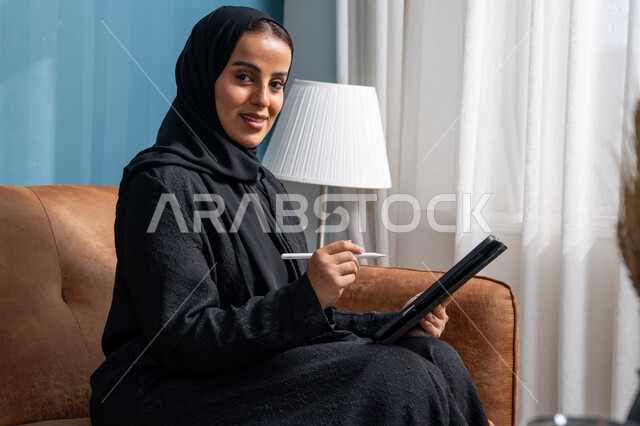 مهنة ووظيفة مكتبية، انجاز وادارة المهام باستخدام جهاز تقني حديث ومتطور, إمرأة عربية خليجية سعودية مبتسمة ترتدي الحجاب والعباءة وتجلس على الكنبة تعمل على جهاز التابلت في صالة المنزل