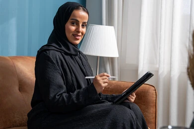 مهنة ووظيفة مكتبية، انجاز وادارة المهام باستخدام جهاز تقني حديث ومتطور, إمرأة عربية خليجية سعودية مبتسمة ترتدي الحجاب والعباءة وتجلس على الكنبة تعمل على جهاز التابلت في صالة المنزل