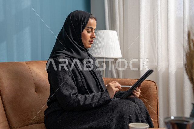 مهنة ووظيفة مكتبية، انجاز وادارة المهام باستخدام جهاز تقني حديث ومتطور, إمرأة عربية خليجية سعودية مبتسمة ترتدي الحجاب والعباءة وتجلس على الكنبة تعمل على جهاز التابلت في صالة المنزل