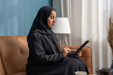 Office profession and job, accomplishing and managing tasks using a modern and advanced technical device, a smiling Saudi Arabian Gulf woman wearing a hijab and abaya sitting on the sofa working on a tablet in the living room