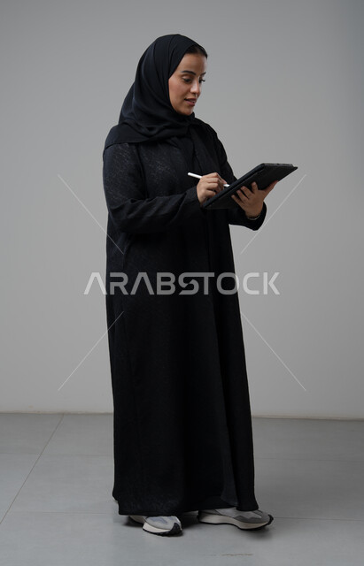 العمل عن بعد، مهنة ووظيفة مكتبية، انجاز وادارة المهام باستخدام جهاز تقني حديث ومتطور, بورتريه لإمرأة عربية خليجية سعودية ترتدي الحجاب والعباءة تعمل على جهاز التابلت، الجسم بالطول الكامل، خلفية رمادية