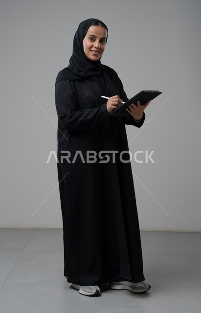 العمل عن بعد، مهنة ووظيفة مكتبية، انجاز وادارة المهام باستخدام جهاز تقني حديث ومتطور, بورتريه لإمرأة عربية خليجية سعودية ترتدي الحجاب والعباءة تعمل على جهاز التابلت، الجسم بالطول الكامل، خلفية رمادية