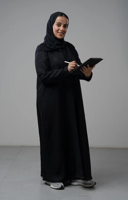 العمل عن بعد، مهنة ووظيفة مكتبية، انجاز وادارة المهام باستخدام جهاز تقني حديث ومتطور, بورتريه لإمرأة عربية خليجية سعودية ترتدي الحجاب والعباءة تعمل على جهاز التابلت، الجسم بالطول الكامل، خلفية رمادية