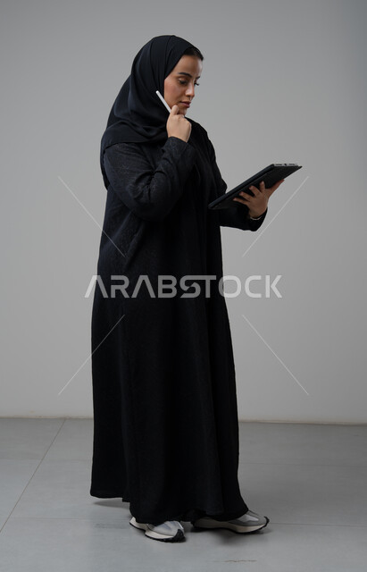 العمل عن بعد، مهنة ووظيفة مكتبية، انجاز وادارة المهام باستخدام جهاز تقني حديث ومتطور, بورتريه لإمرأة عربية خليجية سعودية ترتدي الحجاب والعباءة تعمل على جهاز التابلت، الجسم بالطول الكامل، خلفية رمادية