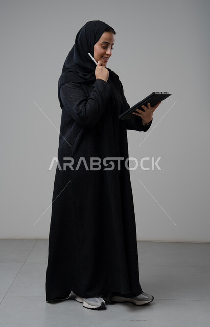 العمل عن بعد، مهنة ووظيفة مكتبية، انجاز وادارة المهام باستخدام جهاز تقني حديث ومتطور, بورتريه لإمرأة عربية خليجية سعودية ترتدي الحجاب والعباءة تعمل على جهاز التابلت، الجسم بالطول الكامل، خلفية رمادية