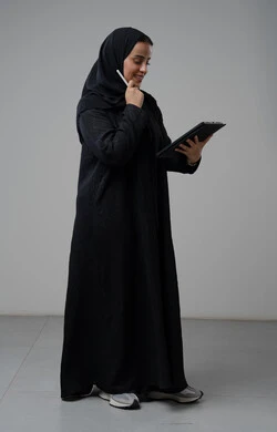 العمل عن بعد، مهنة ووظيفة مكتبية، انجاز وادارة المهام باستخدام جهاز تقني حديث ومتطور, بورتريه لإمرأة عربية خليجية سعودية ترتدي الحجاب والعباءة تعمل على جهاز التابلت، الجسم بالطول الكامل، خلفية رمادية