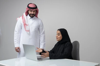 Comfortable work environment in Saudi companies, dialogue and discussion between colleagues, exchange of experiences and information to achieve goals, portrait of two Saudi Gulf Arab employees working in their office inside the company headquarters, use of computers and technology in the business field, gray background