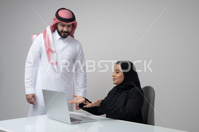 Comfortable work environment in Saudi companies, dialogue and discussion between colleagues, exchange of experiences and information to achieve goals, portrait of two Saudi Gulf Arab employees working in their office inside the company headquarters, use of computers and technology in the business field, gray background