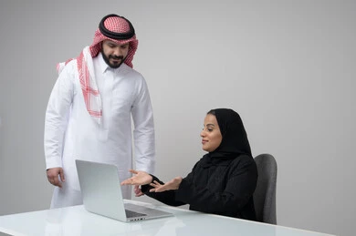 Comfortable work environment in Saudi companies, dialogue and discussion between colleagues, exchange of experiences and information to achieve goals, portrait of two Saudi Gulf Arab employees working in their office inside the company headquarters, use of computers and technology in the business field, gray background