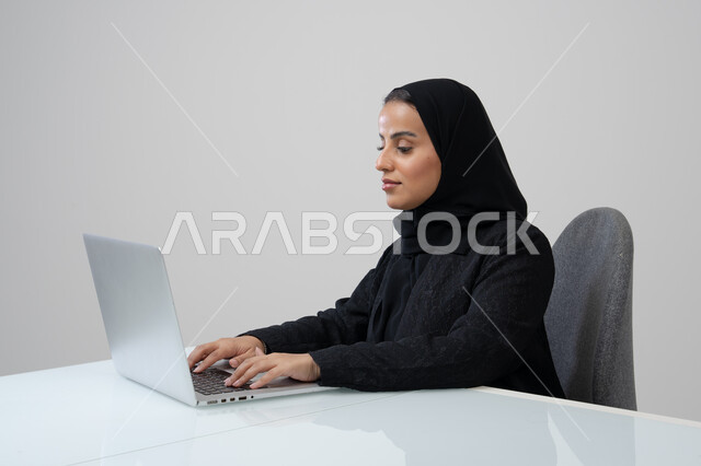 Office job and profession, managing and completing tasks using a modern and advanced technical device, portrait of a Saudi Arabian Gulf woman wearing a hijab and black abaya working on a laptop, gray background