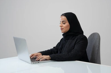 Office job and profession, managing and completing tasks using a modern and advanced technical device, portrait of a Saudi Arabian Gulf woman wearing a hijab and black abaya working on a laptop, gray background