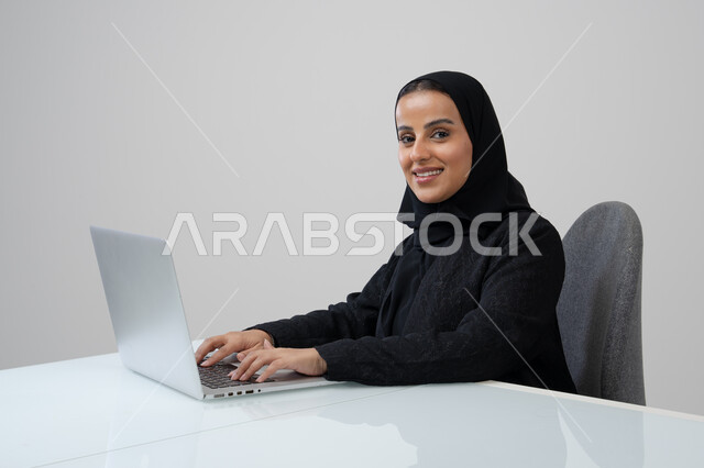 وظيفة ومهنة مكتبية، ادارة وانجاز المهام باستخدام جهاز تقني حديث ومتطور, بورتريه لإمرأة عربية خليجية سعودية ترتدي الحجاب والعباءة السوداء تعمل على الحاسوب المحمول, خلفية رمادية