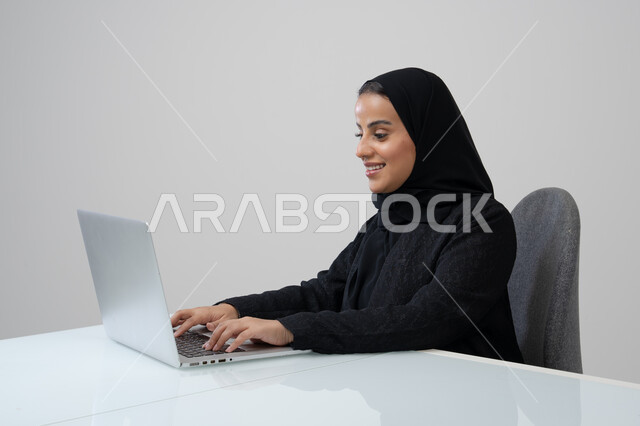 وظيفة ومهنة مكتبية، ادارة وانجاز المهام باستخدام جهاز تقني حديث ومتطور, بورتريه لإمرأة عربية خليجية سعودية ترتدي الحجاب والعباءة السوداء تعمل على الحاسوب المحمول, خلفية رمادية