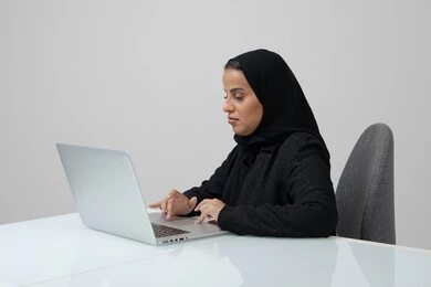 Saudi women's office jobs and professions, managing and completing tasks using a modern and advanced technical device, portrait of a Saudi Arabian Gulf woman wearing a hijab and black abaya working on a laptop at the company headquarters, gray background