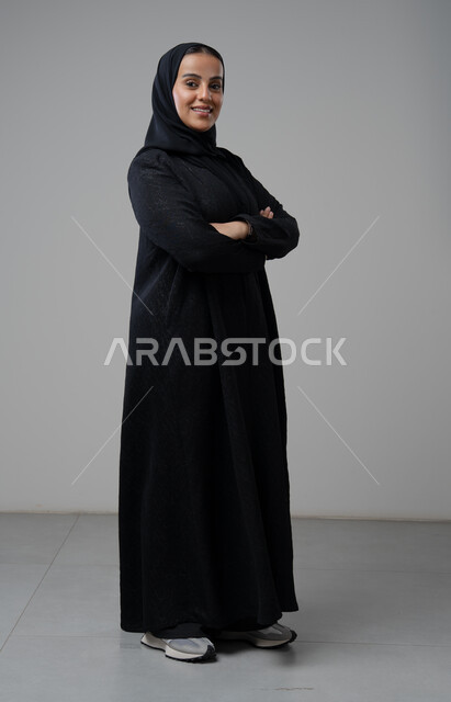 Looking at the camera with gestures of pleasure and self-confidence, the concept of elegance and interest in external appearance, portrait of a Saudi Arabian Gulf woman wearing the hijab and traditional abaya standing with crossed hands, full-length body image, gray background