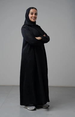 النظر إلى الكاميرا بإيماءات السرور والثقة بالنفس، مفهوم الاناقه و الاهتمام بالمظهر الخارجي، بورتريه لإمرأة عربية خليجية سعودية ترتدي الحجاب والعباءة التقليدية تقف بأيدي متقاطعه، صورة الجسم بالطول الكامل، خلفية رمادية