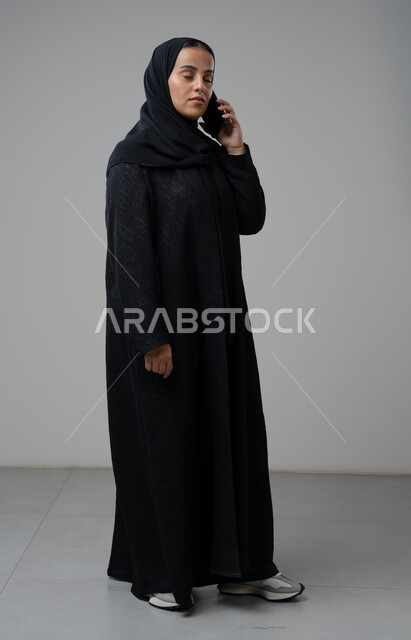 قضاء اوقات ممتعة بتبادل الاخبار والأحاديث، التحدث عبر الهاتف المحمول، بورتريه لامرأة عربية خليجية سعودية محجبة ترتدي العباءة تقوم بإجرء مكالمة هاتفية، محادثات ودردشات مع الاصدقاء، صورة الجسم بالكامل، خلفية رمادية