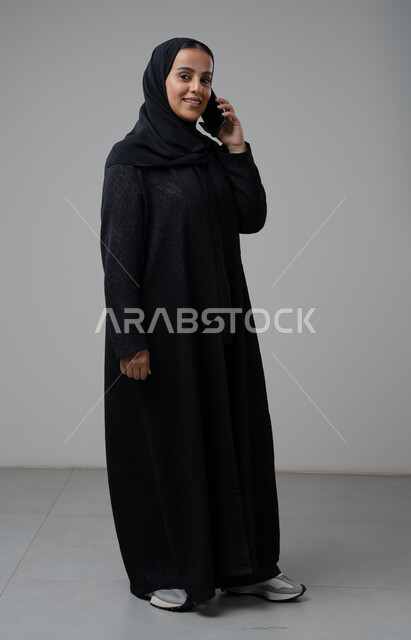 قضاء اوقات ممتعة بتبادل الاخبار والأحاديث، التحدث عبر الهاتف المحمول، بورتريه لامرأة عربية خليجية سعودية محجبة ترتدي العباءة تقوم بإجرء مكالمة هاتفية، محادثات ودردشات مع الاصدقاء، صورة الجسم بالكامل، خلفية رمادية