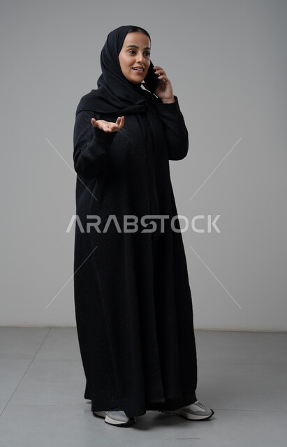 تبادل الاخبار والأحاديث عبر الجوال، التحدث عبر الهاتف المحمول، بورتريه لامرأة عربية خليجية سعودية محجبة ترتدي العباءة ترفع يدها بايماءات التساؤل وتقوم بإجرء مكالمة هاتفية، محادثة ودردشة مع الاصدقاء، صورة الجسم بالكامل، خلفية رمادية