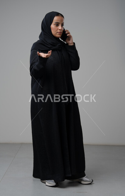 تبادل الاخبار والأحاديث عبر الجوال، التحدث عبر الهاتف المحمول، بورتريه لامرأة عربية خليجية سعودية محجبة ترتدي العباءة ترفع يدها بايماءات التساؤل وتقوم بإجرء مكالمة هاتفية، محادثة ودردشة مع الاصدقاء، صورة الجسم بالكامل، خلفية رمادية