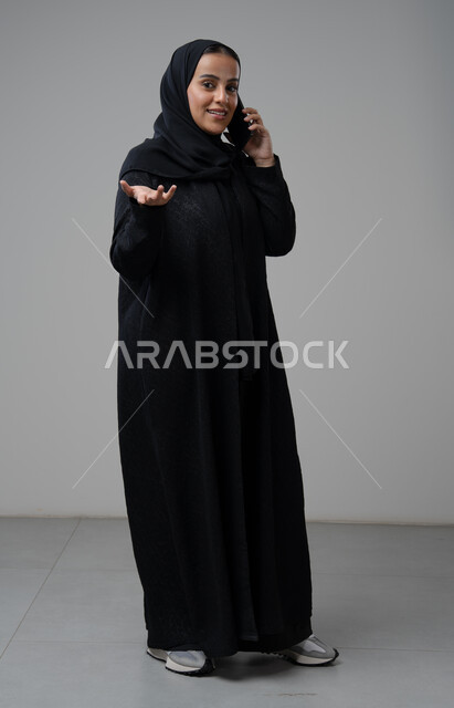 تبادل الاخبار والأحاديث عبر الجوال، التحدث عبر الهاتف المحمول، بورتريه لامرأة عربية خليجية سعودية محجبة ترتدي العباءة ترفع يدها بايماءات التساؤل وتقوم بإجرء مكالمة هاتفية، محادثة ودردشة مع الاصدقاء، صورة الجسم بالكامل، خلفية رمادية
