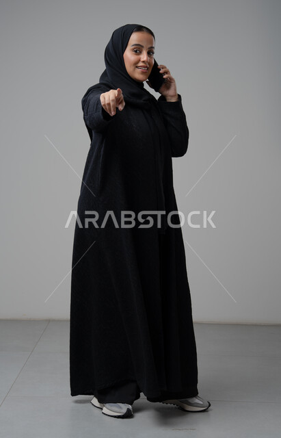 محادثة ودردشة مع الاصدقاء، تبادل الاخبار والأحاديث عبر الجوال، التحدث عبر الهاتف المحمول، بورتريه لامرأة عربية خليجية سعودية محجبة ترتدي العباءة تشير باصبع السبابة إلى شيء ما وتقوم بإجرء مكالمة هاتفية، صورة الجسم بالكامل، خلفية رمادية