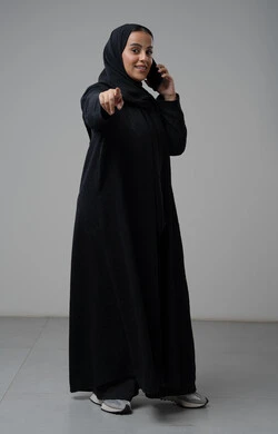 محادثة ودردشة مع الاصدقاء، تبادل الاخبار والأحاديث عبر الجوال، التحدث عبر الهاتف المحمول، بورتريه لامرأة عربية خليجية سعودية محجبة ترتدي العباءة تشير باصبع السبابة إلى شيء ما وتقوم بإجرء مكالمة هاتفية، صورة الجسم بالكامل، خلفية رمادية