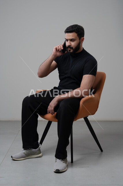 Chatting and conversing using a modern technical device, answering a phone call via mobile phone, portrait of a Saudi Arabian Gulf man in casual clothes sitting on a chair making a phone call via mobile phone, gray background