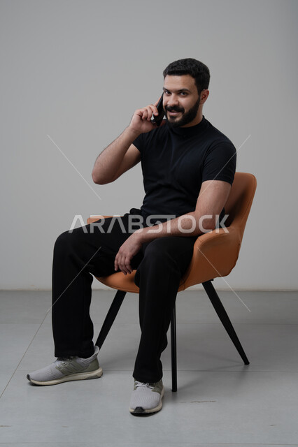 Chatting and conversing using a modern technical device, answering a phone call via mobile phone, portrait of a Saudi Arabian Gulf man in casual clothes sitting on a chair making a phone call via mobile phone, gray background