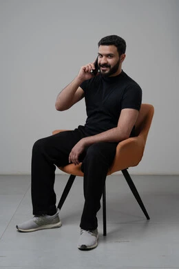 Chatting and conversing using a modern technical device, answering a phone call via mobile phone, portrait of a Saudi Arabian Gulf man in casual clothes sitting on a chair making a phone call via mobile phone, gray background