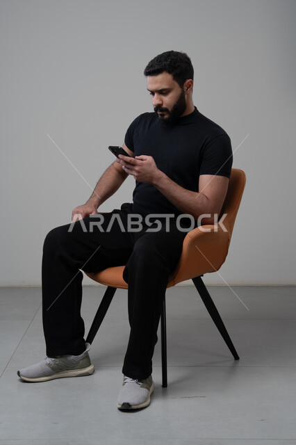 Using modern advanced technology in practical life, working remotely via mobile, gestures of integration and concentration, portrait of a young Saudi Arabian Gulf man wearing casual clothes sitting on a chair working on a mobile phone, gray background
