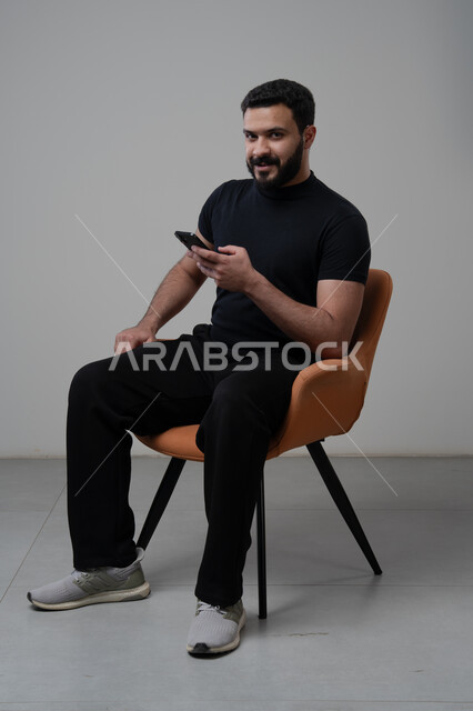 استخدام التقنية الحديثه المتطورة في الحياة العملية، العمل عن بعد عبر الجوال، ايماءات الاندماج والتركيز، بورتريه لشاب عربي خليجي سعودي يرتدي الزي الكاجوال يجلس على الكرسي يقوم بالعمل على الهاتف المحمول، خلفية رمادية