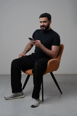 استخدام التقنية الحديثه المتطورة في الحياة العملية، العمل عن بعد عبر الجوال، ايماءات الاندماج والتركيز، بورتريه لشاب عربي خليجي سعودي يرتدي الزي الكاجوال يجلس على الكرسي يقوم بالعمل على الهاتف المحمول، خلفية رمادية