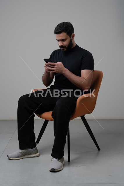Using modern advanced technology in practical life, working remotely via mobile, gestures of integration and concentration, portrait of a young Saudi Arabian Gulf man wearing casual clothes sitting on a chair working on a mobile phone, gray background
