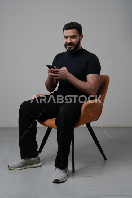 Using modern advanced technology in practical life, working remotely via mobile, gestures of integration and concentration, portrait of a young Saudi Arabian Gulf man wearing casual clothes sitting on a chair working on a mobile phone, gray background